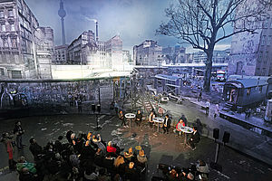 Auf dem Bild sieht man ein sitzendes Publikum auf eine Gruppe Menschen schauen, die ein Podium darstellen und miteinander diskutieren. Hinter ihnen ist ein 360° Panorama von der Berliner Mauer zu sehen.