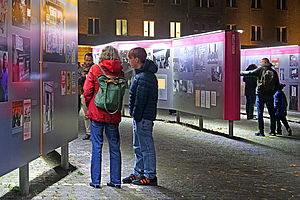 Auf dem Bild sind mehrere Personen zu sehen, die sich die Open-Air-Ausstellung nachts auf dem Campus für Demokratie anschauen.