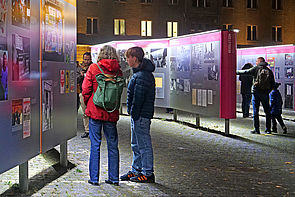 Auf dem Bild sind mehrere Personen zu sehen, die sich die Open-Air-Ausstellung nachts auf dem Campus für Demokratie anschauen.
