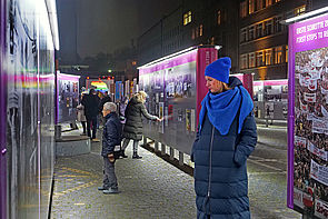 Auf dem Bild ist eine Frau zu sehen, die sich die Open-Air-Ausstellung auf dem Campus für Demokratie anschaut. Man sieht, dass es nachts ist. Die Ausstellung ist beleuchtet.