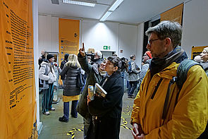 Auf dem Bild sind mehrere Menschen in einem Ausstellungsraum zu sehen, die sich die Texte und Objekte an den Wänden finden, anschauen.