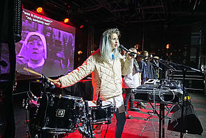 Auf dem Bild sieht man eine junge Frau, die auf einer Bühne neben weiteren Künstlerinnen steht und in ein Mikrofon singt. Daneben stehen diverse elektronische Geräte und Musikinstrumente. Im Hintergrund ist ein projiziertes Bild zu sehen.