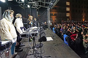 Auf dem Bild sieht man eine junge Frau, die auf einer Bühne neben weiteren Künstlerinnen steht und in ein Mikrofon singt. Daneben stehen diverse elektronische Geräte und Musikinstrumente. Im Hintergrund ist ein projiziertes Bild zu sehen.