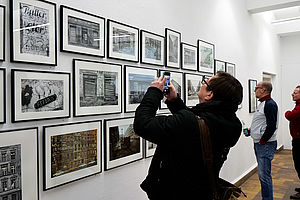 Auf dem Bild sieht man in der Mitte einen Mann in dunkler Kleidung, der mit seinem Handy ein Foto der vor ihm an der Wand befindlichen Bilder macht. Im Hintergrund sieht man zwei weitere Männer, die sich die Fotoausstellung anschauen.