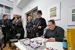 Auf dem Bild sieht man einen Mann an einem Tisch sitzen, der vor ihm liegende Bücher signiert. Links daneben sieht man weitere Personen, die in dem Raum stehen.