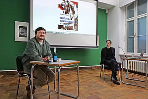 Auf dem Bild sieht man zwei Personen. Links sitzt ein Mann an einem kleinen Tisch. Hinter ihm ist eine Leinwand zu sehen, an die ein Buchcover projiziert ist.