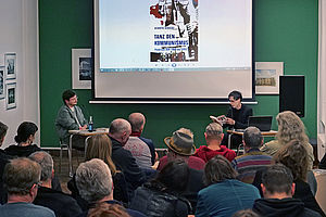 Auf dem Bild sieht man zwei Menschen, die auf einer Art Podium sitzen vor einem sitzen Publikum, das in ihre Richtung blickt.
