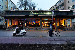 Auf dem Bild ist eine Gebäude mit großen Glasflächen zu sehen. Es handelt sich dabei um eine Bar, die in Berlin-Prenzlauer Berg steht. Der Innenbereich ist mit gelblich-warmen Licht beleuchtet. Ein Leuchtschild befindet sich darüber, das den Schriftzug "Supersonico" trägt.