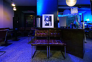 Auf dem Bild sieht man ein Schwarz-Weiß-Foto, das auf einem Tisch steht und den jungen Olaf Weißbach zeigt. Das Licht in der Bar, in der sich der Tisch mit den zwei davor befindlichen braunen Lederstühlen befindet, leuchtet blau.