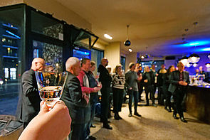 Auf dem Bild sieht man eine Gruppe Menschen in einer Bar stehend und mit einem Glas in der Hand. Auf der linken Seite sieht man eine Hand, die ein Glas in der Hand hält und zum Prosten in die Höhe reckt.