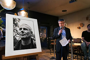 Auf dem Bild sieht man einen Mann, der ein Mikrofon in der rechten Hand hält und in der linken Hand ein Blatt Papier. Auf der linken Seite des Bildes ist eine Staffelei zu sehen, auf der eine Schwarz-Weiß-Fotografie von Olaf Weißbach zu sehen ist.