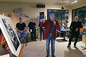 Auf dem Bild ist ein Man mit rotem Jackett zu sehen, der ein Mikrofon in der Hand hält. Auf der rechten Seite sieht man eine schwarz-weiß-Fotografie von Olaf Weißbach auf einer Staffelei stehen. Im Hintergrund sieht man weitere Personen zum Teil stehend zum Teil sitzend, die zuhören.