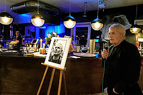 Auf dem Bild sieht man Robert Jahn, der ein Mikrofon in der Hand hält. Neben ihm steht eine Staffelei mit einem Schwarz-weiß-Foto von Olaf Weißbach. Im Hintergrund sieht man eine Bar mit einem Bartresen, über dem sich hängende runde Lampen befinden.