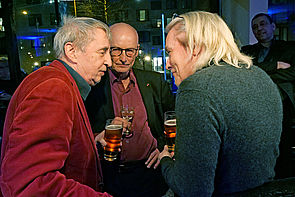 Auf dem Bild sieht man drei Männer, die sich intensiv unterhalten. Zwei von ihnen halten ein Glas Bier in der Hand.