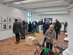 Auf dem Bild sieht man mehrere Menschen in einem Galerieraum, die sich die an den Wänden befindenden aufgehängten und gerahmten Fotografien anschauen.