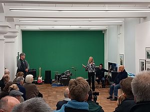 Auf dem Bild sieht man im Vordergrund die Hinterköpfe von mehreren Menschen. Alle blicken auf eine grün gestrichene Wand, vor der eine Frau am Mikrofon steht und zu den Personen spricht.