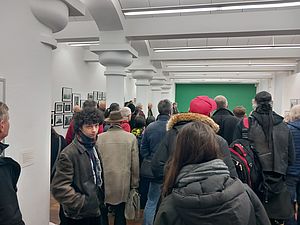 Auf dem Bild sieht man eine große Menschenmenge, die sich dicht gedrängt in einem Galerieraum befindet und in die Richtung einer grün gestrichenen Wand blickt.