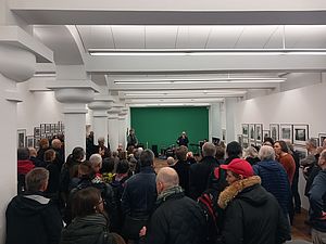 Auf dem Bild sieht man eine große Gruppe Menschen, die in die gleiche Richtung auf eine grün gestrichene Wand blicken, vor der ein kleines Podium aufgebaut ist.
