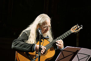 Auf dem Bild ist eine älterer Mann zu sehen, der auf einer Gitarre spielt. Er hat lange weiße Haare. Vor ihm steht ein Notenständer und ein Mikrofonständer.