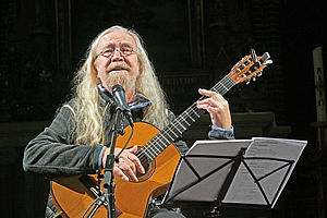 Auf dem Bild ist ein Mann mit weißen langen Haaren zu sehen, der eine Gitarre in seiner Hand hält. Vor ihm steht ein Notenständer und ein Mikrofon.