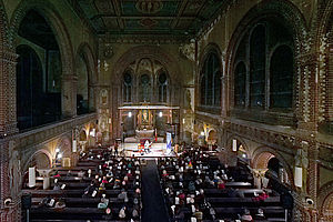 Auf dem Bild sind Menschen in einem Kirchenschiff zu sehen, die auf Bänken sitzen und auf eine Bühne mit einem Musiker schauen.