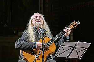 Auf dem Bild ist ein Mann mit langen weißen Haaren zu sehen, der eine Gitarre in der Hand hält. Er singt in ein Mikrofon. Vor ihm steht ein Notenständer.