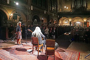 Auf dem Bild ist ein Mann auf der Bühne sitzend zu sehen. Der Gitarrenspieler sitzt auf einem Stuhl und schaut in ein Publikum. Er ist nur von hinten zu sehen und hat lange weiße Haare.
