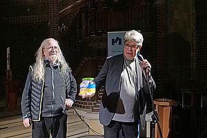 Auf dem Bild ist ein Mann mit weißen langen Haaren und eine Frau an einem Mikrofon stehend zu sehen.