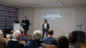 Auf dem Bild sieht man einen Referenten und eine Referentin, die in dunkler Kleidung auf einem Podium stehen und vor einer mit einem Beamer an die Wand geworfener Präsentation stehen. Die Frau hält einen Zettel in der Hand, spricht zum Publikum und stellt den Referenten, neben ihr stehend, vor. Davor sitzen Menschen auf Stühlen und blicken in die Richtung des Vortragenden.