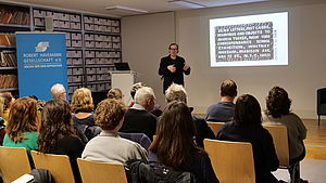 Auf dem Bild sieht man einen Referenten in dunkler Kleidung auf einem Podium stehen, der vor einer mit einem Beamer an die Wand geworfener Präsentation steht. Davor sitzen Menschen auf Stühlen und blicken in die Richtung des Vortragenden.
