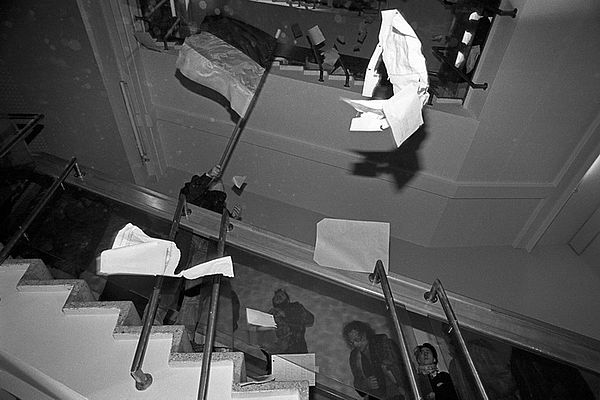 Auf dem schwarz-weiß Foto sieht man ein Treppenhaus, das von unten nach oben fotografiert wurde. Es fliegen weiße Papierblätter durch die offene Mitte des Treppenhauses. Auf den Treppen befinden sich mehrere Menschen, die zum Teil nach oben oder nach unten laufen. Eine Person, die auf dem Weg nach unten ist, trägt eine Deutschlandflagge in der Hand.