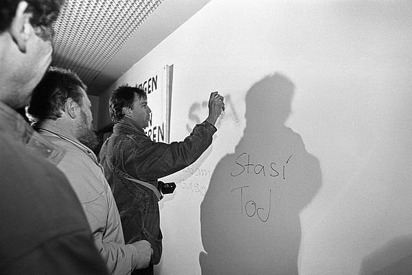 Auf dem schwarz-weiß Foto sieht man drei Männer, die vor einer weißen Wand in einem Gebäude, vermutlich in einem Gebäude auf dem Areal der Stasi-Zentrale in Berlin-Lichtenberg, stehen. Im Vordergrund sieht man den seitlichen Anschnitt zweier Männer. In der Bildmitte steht ein Mann, den man von der Seite sieht. Er hält etwas in seiner rechten Hand, womit er ein Wort an die Wand sprüht. Auf der Wand kann man bereits die Wörter "Stasi Tod", "15.1.1990" und die Großbuchstaben STA lesen. Der Mann in der Mitte des Bildes wirft einen Schatten an die weiße Wand, womit seine Umrisse gut zu erkennen sind.