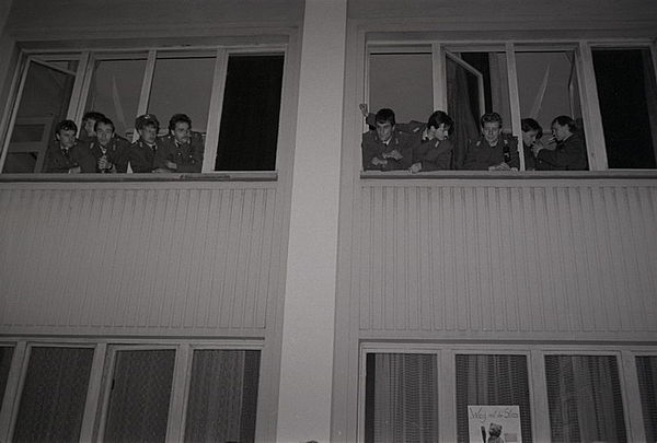 Auf dem schwarz-weiß Foto sind neun junge Männer in Uniform zu sehen, die sich aus zwei geöffneten Fenstern eines Gebäudes nach draußen beugen und in die Umgebung schauen. Unter den geöffneten Fenstern befindet sich eine weitere Etage, ebenfalls mit Fenstern.