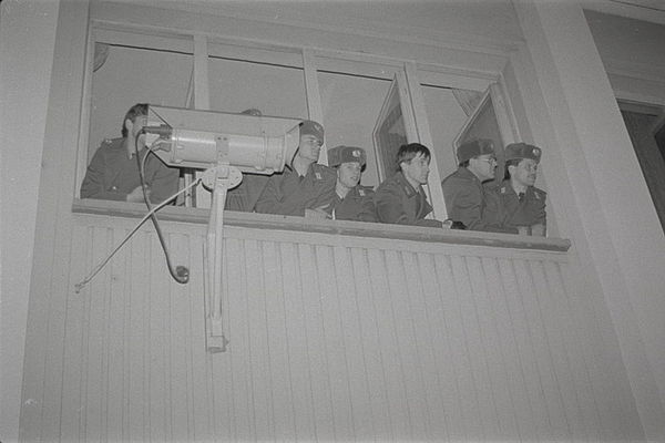 Auf dem Bild sieht man sieben Männer in Uniform aus mehreren geöffneten Fenstern nach draußen schauend. Unterhalb des geöffneten Fenster ist links auch eine Überwachungskamera angebracht an der Außenhaut des Gebäudes.