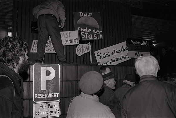 Auf dem schwarz-weiß Foto sieht man Menschen von hin. Man sieht zudem eine Vielzahl an Transparenten und Demonstrationsschildern. Ein Mann steht au einer Mauer, auf dem die Transparente und Demonstrationsschilder abgestellt sind. An der Mauer weiter unten ist ein Parkplatzschild angebracht.