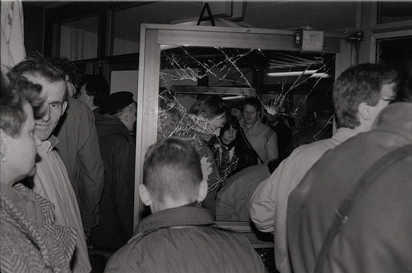Auf dem schwarz-weiß Foto sieht man mehrere Menschen, die gerade dabei sind durch eine kaputte Glastür zu gehen.