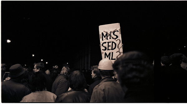Auf dem schwarz-weiß Foto sieht man eine Menschenmenge, die in eine Gruppe läuft. Auf dem Bild sieht man die man mittig ein Transparent, das über die Köpfe der Menschenmenge hinausragt. Auf dem Transparent steht "MfS, SED?, ML?". Das Wort "MfS" wurde dabei durchgestrichen.