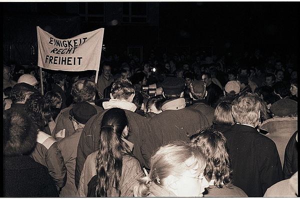 Auf dem schwarz-weiß Foto ist eine größere Menschenmenge zu sehen, die in eine Richtung läuft. Dias Foto zeigt die Menschengruppe von hinten. Am linken Bildrand sieht man ein Transparent sieht man ein Transparent, das aus der Menschenmenge ragt. Auf dem Transparent steht "Einigkeit, Recht, Freiheit".