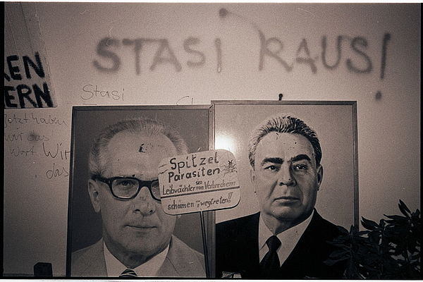 Auf der schwarz-weiß Fotografie sieht man eine weiße Innenwand, die mit Wörtern beschmiert ist und an der zwei gleichgroße Bilder in Bilderrahmen lehnen. Vor den Bilderrahmen ist ein kleines Demonstrationsschild zu sehen, das aus einer rechteckigen Pappe mit abgerundeten Ecken besteht, das an einem Stab befestigt ist. Auf dem Schild steht "Spitzel Parasiten, SED Leibwächter von Verbrechern, schämen, wegtreten!". In dem rechten Bilderrahmen ist ein Foto von Leonid Iljitsch Breschnew zu sehen. In dem linken Bilderrahmen befindet sich ein Foto von Erich Honecker. Auf der Wand dahinter stehen geschrieben die Worte "Stasi raus!", "Stasi" und "Jetzt haben wir das Wort".