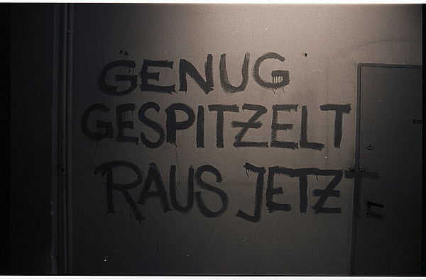 Auf dem schwarz-weiß Foto ist eine weiß getünchte Innenwand zu sehen, auf der in Großbuchstaben zu lesen ist "Genug gespitzelt raus jetzt". Auf der linken Seite der Wand verlaufen mehrere weiße Rohre, auf der rechten Seite ist eine graue Tür zu sehen.