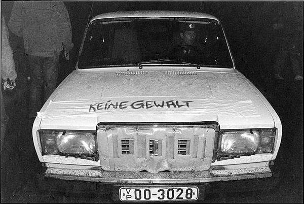 Auf dem schwarz-weiß Bild ist ein Auto frontal von vorn zu sehen. In dem Fahrzeug sitzt ein Mann am Lenkrad. Man sieht ebenso das Nummernschild unterhalb des Kühlergrills des Fahrzeuges. Auf der linken Seite sieht man Menschen, die in die Gegenrichtung des Autos laufen.