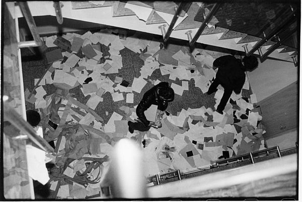 Auf dem schwarz-weiß Foto sieht man ein Treppenhaus aus der Perspektive von oben fotografiert. Auf dem Boden, dem Ende des Treppenhauses, liegen viele lose Blatt Papier. Zwei dunkel gekleidete Männer laufen darauf herum und schauen dabei auf den Boden.