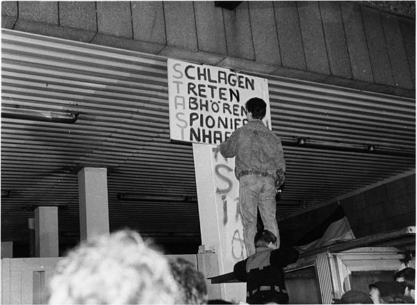 Auf dem schwarz-weiß Foto ist ein junger Mann zu sehen, der auf dem Dach eines Wachhauses steht und gerade dabei ist, ein Transparent an eine Säule anzuschlagen. Auf dem Transparent stehen die Worte "Schlagen, Treten, abhören, spionieren, inhaftieren". Ein weiterer junger Mann ist gerade dabei, sich aus eigener Kraft auf das Dach des Wachhauses zu ziehen.