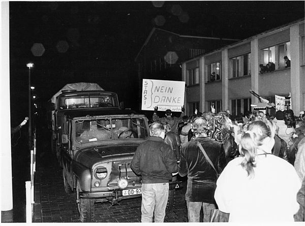 Auf dem schwarz-weiß Foto ist ein Geländewagen zu sehen, der auf dem Gelände der Stasi-Zentrale in Berlin-Lichtenberg steht. Rechts an dem Wagen vorbei läuft eine Menschenmenge. Darunter sind Männer als auch Frauen. Eine Person aus der Menschenmenge hält ein Transparent hoch auf dem die Worte "Stasi, nein danke" steht.