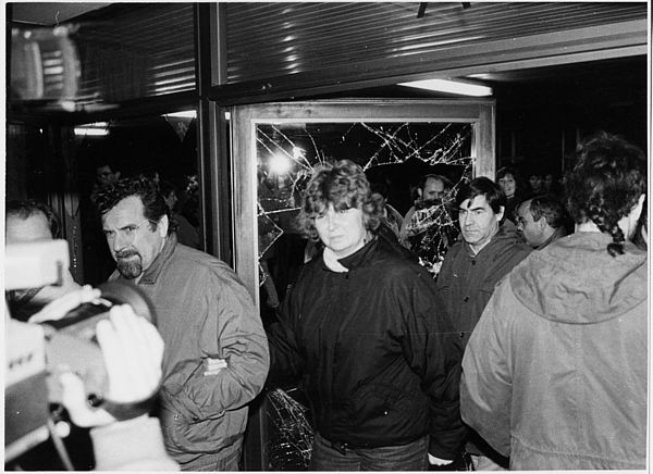Auf dem schwarz-weiß Foto sieht man einen Mann und eine Frau im Zentrum des Bildes, die gerade ein Gebäude betreten, das durch eine kaputte Eingangstür mit zerstörter Glasscheibe, hineingehen. Hinter der Frau und dem Mann, die direkt in die Kamera des Fotografen blicken, läuft ein weiterer Mann. Im Hintergrund sieht man eine weiter Menschengruppe, die dabei ist, das Gebäude zu betreten. Am linken Rand des Bildes ist die Hand eines Menschen an einer Filmkamera zu sehen, die ebenfalls die Szenerie festhält.