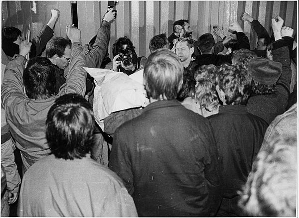 Auf dem schwarz-weiß Foto sieht man eine Gruppe von Menschen, die, so scheint es, sich um eine Person herum versammelt haben. Menschen im hinteren linken Bereich des Fotos haben Kameras in der Hand, die sie mit einer Hand in die Höhe strecken. um von oben ein Foto zu machen. Die anderen Menschen im Vordergrund des Bildes sind nur von hinten zu sehen. Die Person auf die sich der Fokus der Fotografen richtet, hat eine Art weiße Plane über dem Oberkörper. Einige Menschen auf der rechten Seite des Bildes, die drum herum stehen, recken ihre rechten Fäuste in die Luft.