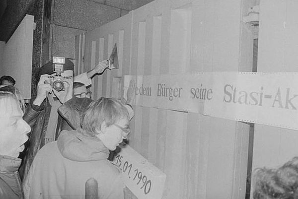 Auf dem Bild sieht man Menschen, die vor einem Tor stehen. Darauf kleben Streifen, auf denen sich Schrift befindet.
