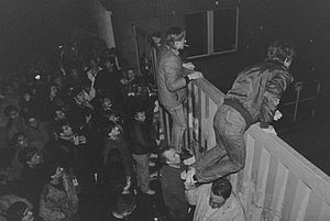 Auf der schwarz-weiß-Fotografie sieht man im Vordergrund zwei Männer, die dabei sind, ein hohes Tor mit eigener Muskelkraft zu überwinden. Hinter ihnen steht eine große Menschenmenge.