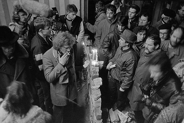 Auf der schwarz-weiß Fotografie sind Menschen um eine kleine Ziegelmauer herum versammelt, die die Menschen symbolisch bei der Erstürmung der Stasi-Zentrale in Berlin spontan errichtet haben.