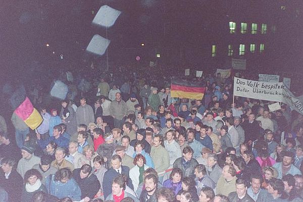 Auf dem Bild sieht man eine große, dicht gedrängte Menschenmenge, die auf das Gelände der Stasi-Zentrale stürmen. Einige Davon halten Transparente oder Deutschlandfahnen in die Höhe.